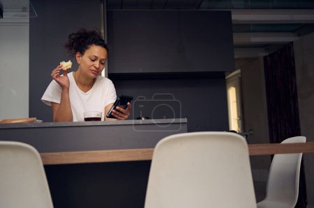 Femme multi ethnique confiante 40 ans, utilisant un smartphone, vérifiant le contenu des médias sociaux, naviguant sur le web, faisant défiler les newsfeed, debout au comptoir de la cuisine avec un sandwich à la main, profitant de son petit déjeuner à la maison
