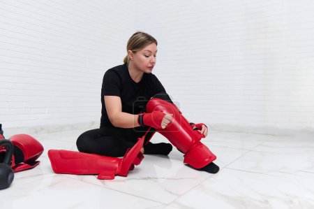 Full size shot of European young woman 30s, boxer fighter putting on red kickboxing leg pads guard. Sparring shin guards. Martial art, combat, challenge. Kickboxing concept. People, sport and fitness