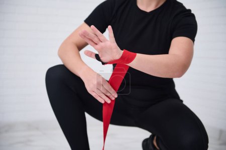 Peleadora joven fuerte de primer plano envolviendo su muñeca y manos con una cinta roja, preparándose para el entrenamiento de boxeo, aislada sobre fondo blanco. Deporte, ejercicio cardiovascular, resistencia y resistencia