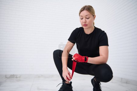 Caucasian young woman boxer fighter preparing boxing bandages, wrapping her wrist and hands with a red tape, before putting on boxing gloves, getting ready for training, isolated over white background