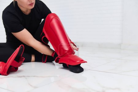 Enfoque selectivo en las piernas de una boxeadora mujer que se pone almohadillas rojas para las piernas. Guardias de espinilleras. Arte marcial, combate, desafío. Concepto de Kickboxing. Personas, deporte y fitness