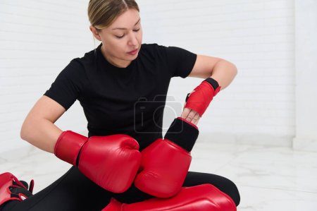 Young Caucasian woman fighter, boxer putting on red boxing gloves over white wall background