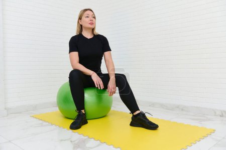 Una joven deportista haciendo ejercicio en un estudio en casa usando equipo de gimnasia. Ella está sentada en una pelota de ejercicio verde, vestida con ropa deportiva negra, en una alfombra de ejercicio amarilla en un entorno minimalista.