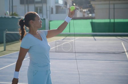 Mujer de mediana edad practicando tenis en una cancha al aire libre. Ella está sosteniendo la pelota de tenis con expresión enfocada, usando atuendo deportivo y accesorios de tenis, destacando la dedicación al fitness y los deportes.