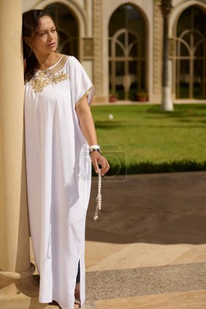 Elegant woman in a beautiful white traditional dress, adorned with golden embroidery, standing by a column outdoors. She appears thoughtful and serene, holding a string of prayer beads.