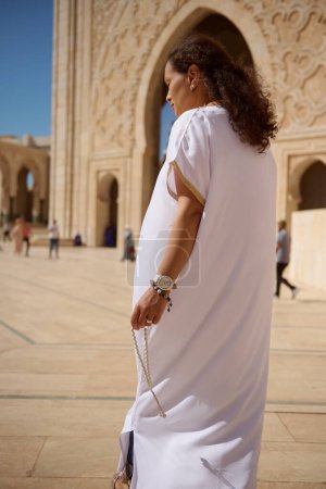 Une femme en costume blanc traditionnel tient des perles de prière et se tient devant grand une entrée de mosquée ornée. Une architecture complexe et une atmosphère lumineuse indiquent un moment spirituel serein.