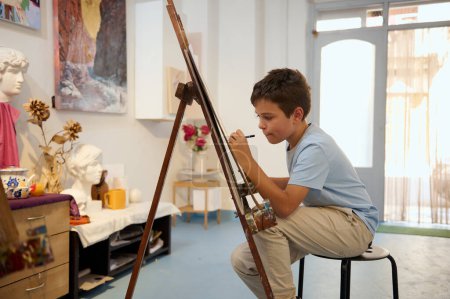 Ein kleiner Junge im Kunstunterricht, der sich auf Zeichnen und Malen an einer Staffelei in einer gut ausgestatteten Werkstatt konzentriert. Kreatives und pädagogisches Umfeld.