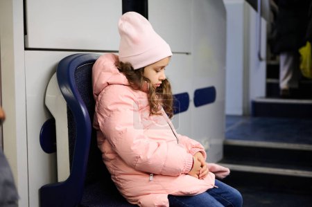 A young girl wearing a pink jacket and hat sits alone on a train, looking thoughtful and introspective. The image captures the mood of solitude and contemplation during travel.
