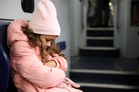 Ein junges Mädchen in rosa Mantel und Hut sitzt allein auf einem Zugsitz und blickt nach unten und vermittelt Gefühle der Einsamkeit und Kontemplation.