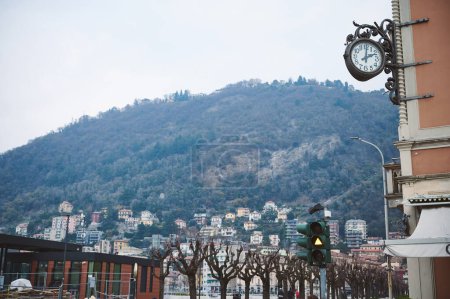 Stadtbild mit einer traditionellen Uhr an einem Gebäude, mit Blick auf saftig grüne Berge, kahle Bäume und Wohnhäuser, die über den Hang verstreut sind.