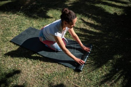 Woman rolling up a yoga mat on grass in a sunny outdoor park. Relaxation, fitness, and mindfulness in a serene natural setting.