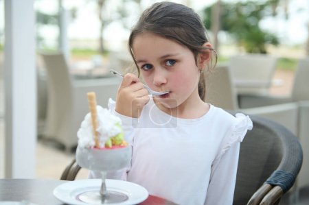 Chica joven con camisa blanca saboreando helado en la zona de estar al aire libre de un café. Ella se ve contenta y concentrada.