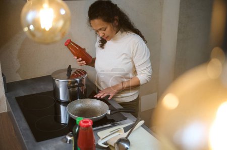 Woman cooking in a modern kitchen, holding a bottle of sauce and checking cookware on an electric stove. Warm lighting adds ambiance.