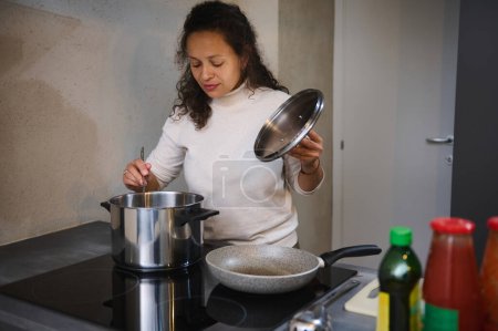 Woman in a modern kitchen stirring ingredients in a pot while holding the lid. Various kitchen items are visible on the countertop.