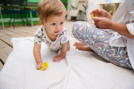 Adorable bebé arrastrándose sobre una manta con un juguete amarillo, acompañado por una mujer en traje colorido. Escena al aire libre con un sentido de exploración y vinculación.