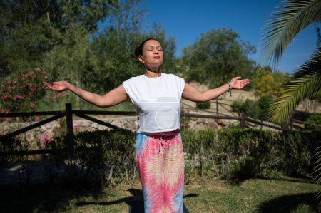 A woman meditates outdoors in a tranquil garden, enjoying the peace and warmth of the sun. She stands with closed eyes and outstretched arms, embracing serenity and mindfulness.