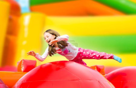 Téléchargez les photos : Joyeux petite fille ayant beaucoup de plaisir sur un château gonflable tout en sautant. Aire de jeux colorée. - en image libre de droit