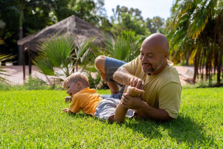 family and love concept - young father tickling his little son in park. dad tickles his son's legs on the grass. dad with little son having fun on the green lawn