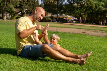 concept de famille et d'amour - le jeune père chatouille son petit fils dans le parc. Papa chatouille les jambes de son fils sur l'herbe. papa avec petit fils s'amuser sur la pelouse verte