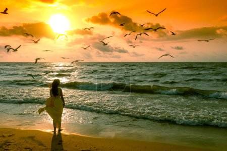 Foto de Paisaje marino durante el amanecer. Mujer en la playa. Una joven mujer feliz con un vestido ondeante camina a lo largo de la orilla del mar - Imagen libre de derechos