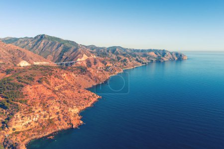 Rocky seashore with clear blue sky. Seascape in the evening. Aerial view from Maro Beach to Pigeon Cave and Cerro Gordo viewpoint. La Miel Viaduct on Mediterraneo Highway. Malaga, Granada, Spain