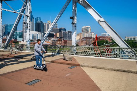 Téléchargez les photos : Nashville, Tennessee États-Unis - 12 mai 2023 : Vue sur les toits du centre-ville depuis le pont piétonnier John Seigenthaler vintage au-dessus de la rivière Cumberland avec un cycliste de scooter passant par - en image libre de droit