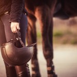 Equestrian girl standing next to her horse and holding her equestrian helmet. Equestrian sport theme.