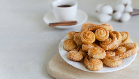 Puff pastry cookies with sugar on a white plate with a small white cup with coffee and a cinnamon stick. The concept of homemade baking. copy space Horizontal orientation. Selective focus