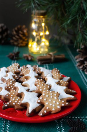 Festliche Plätzchen mit weißem Zuckerguss auf rotem Teller vor grünem Hintergrund mit Lichtern. Hausgemachte Ingwer-Zimt-Weihnachtsplätzchen. Vertikale Orientierung. Selektiver Fokus.