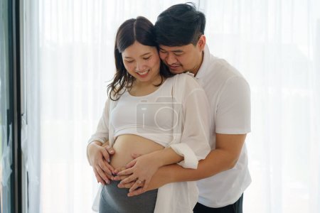 Photo for Happy Asian couple expecting baby standing together against window at home, loving husband tenderly touching belly of his pregnant wife - Royalty Free Image