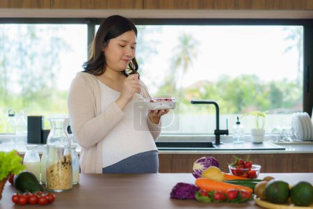 Téléchargez les photos : Beautiful Asian pregnant woman eating with lust cereals on breakfast in kitchen at home, enjoying meal. Yummy food for pregnant - en image libre de droit
