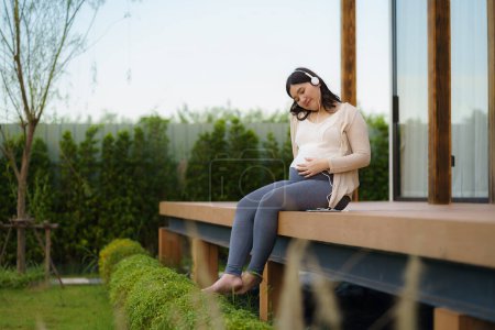 Téléchargez les photos : Portrait of pregnant Happy Asian woman sitting on terrace at home and using smart phone for listening music in headphones. - en image libre de droit