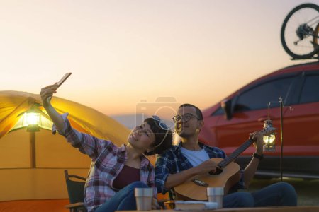 Téléchargez les photos : Asian couple singing and playing guitar while taking a selfie by smartphone camera in their camping area with lake in the background during sunset. - en image libre de droit