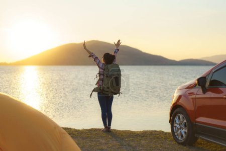 Téléchargez les photos : Asian backpacker woman raising her hands happy to arrive at her destination where she is camping with a lake in the background during sunset. - en image libre de droit