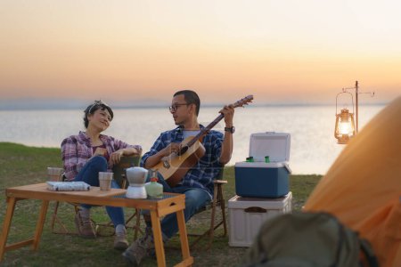Téléchargez les photos : Asian couple singing and playing guitar in their camping area with lake in the background during sunset. - en image libre de droit