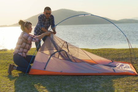 Téléchargez les photos : Asian couple preparing a tent to camping in the lawn with the lake in the background during sunset - en image libre de droit