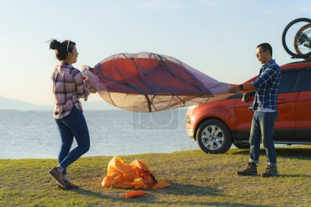 Téléchargez les photos : Asian couple preparing a tent to camping in the lawn with the lake in the background during sunset - en image libre de droit