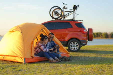 Téléchargez les photos : Young Asian couple taking selfie by smartphone camera by sitting in a tent while camping with the lake in the background during sunset - en image libre de droit