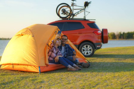 Téléchargez les photos : Young Asian couple taking selfie by smartphone camera by sitting in a tent while camping with the lake in the background during sunset - en image libre de droit
