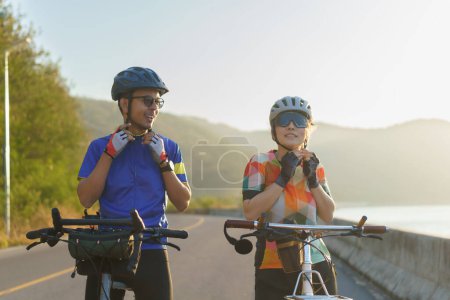 Téléchargez les photos : Asian cyclist couple dons helmets and chin straps as they prepare for their bike ride around a beautiful morning lake. - en image libre de droit