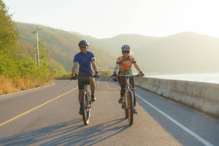 Téléchargez les photos : Couple cycliste asiatique chevauchant ensemble pour l'exercice autour du lac le matin avec belle vue sur la montagne en arrière-plan. - en image libre de droit