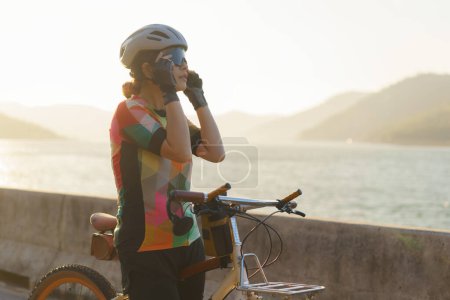 Téléchargez les photos : Femme cycliste asiatique portant des lunettes de soleil se préparer pour une balade à vélo matinale autour du lac avec belle vue sur la montagne en arrière-plan. - en image libre de droit