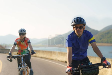 Téléchargez les photos : Couple cycliste asiatique chevauchant ensemble pour l'exercice autour du lac le matin avec belle vue sur la montagne en arrière-plan. - en image libre de droit