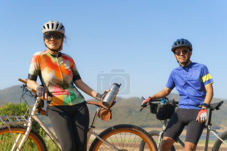 Téléchargez les photos : Tired Asian cyclist couple taking a break to drink water from a bottle with a beautiful lake and mountains in the background. - en image libre de droit