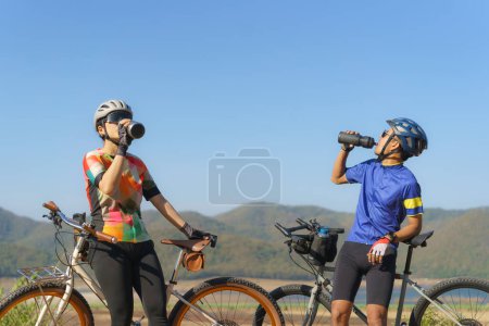 Téléchargez les photos : Tired Asian cyclist couple taking a break to drink water from a bottle with a beautiful lake and mountains in the background. - en image libre de droit