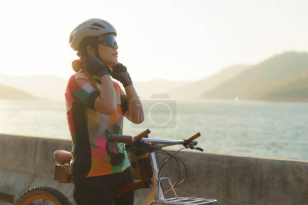 Téléchargez les photos : Asian woman cyclist wearing a helmet getting ready for a bike ride around the lake in the morning with beautiful mountain view in the background. - en image libre de droit