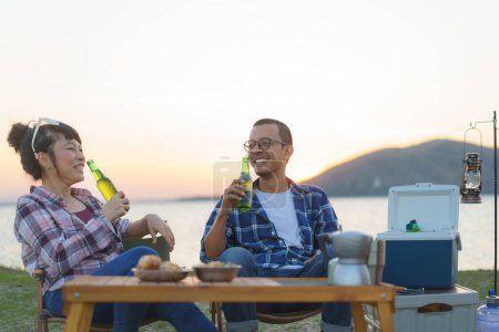 Téléchargez les photos : Couple asiatique boire de la bière de la bouteille dans leur zone de camping avec lac en arrière-plan pendant le coucher du soleil. - en image libre de droit