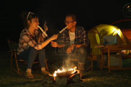 Téléchargez les photos : Asian couple roasting marshmallows together at a campfire where they set up a tent to camp by the lake at night. - en image libre de droit