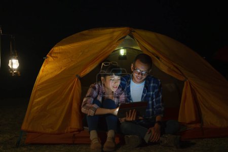 Téléchargez les photos : Asian couple is playing on their digital tablet in his tent during the night planning a trip for tomorrow. - en image libre de droit