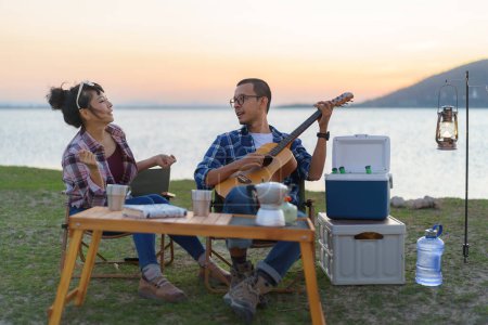 Téléchargez les photos : Asian couple singing and playing guitar in their camping area with lake in the background during sunset. - en image libre de droit